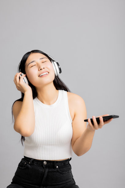 高兴地松了一口气的亚洲日本女人，长黑发，闭着眼睛，在手机里唱歌，就像麦克风一样，戴着现代的新耳机，在灰色的背景下听音乐图片下载