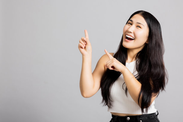 迷人的年轻亚洲女孩女士，长黑发，看着相机微笑，张开嘴好奇地指着下面的东西，她的健康护理护肤图片下载