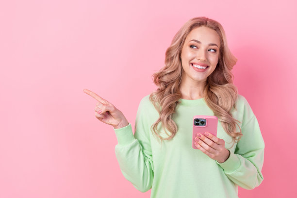 年轻女孩的照片指着空白的空间推荐购物网站亚马逊提供广告持有iphone孤立的粉红色背景图片下载