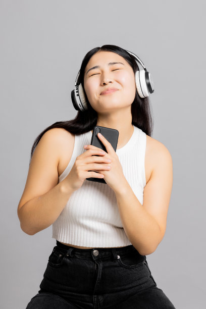 高兴地松了一口气的亚洲日本女人，长黑发，闭着眼睛，在手机里唱歌，就像麦克风一样，戴着现代的新耳机，在灰色的背景下听音乐图片下载
