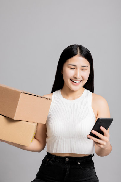 惊讶的年轻亚洲、日本、韩国青少年博主，手里拿着装满物品的盒子，拿着手机自拍，看着手机里不同的照片，站在灰色的背景上，孤立地站着图片下载