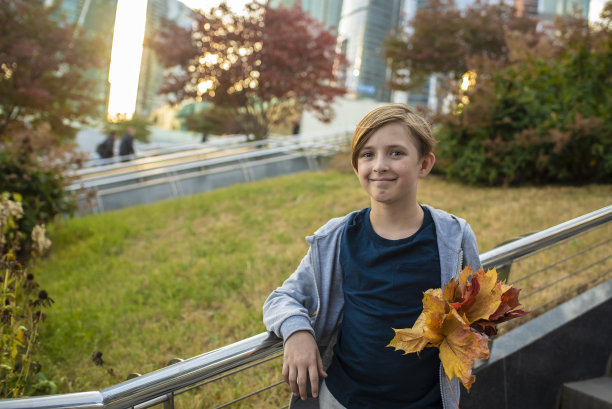 一个11岁的男孩背着背包，手里拿着秋天的枫叶和黄叶，面带微笑。学生，在摩天大楼的背景下的孩子，大都市在秋天上学。图片下载