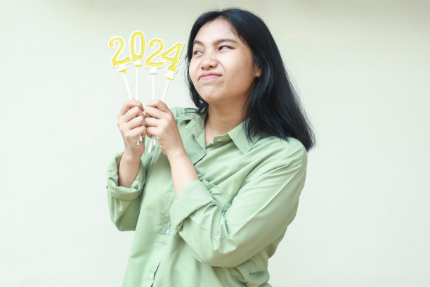 快乐的亚洲年轻女子一边看着闪闪发光的2024号蜡烛，想着新年庆祝的决心，穿着白色背景的绿色衬衫图片下载