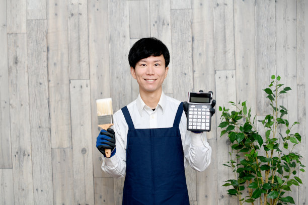 一个面带微笑的年轻亚洲人，系着围裙，手里拿着画笔和计算器图片下载