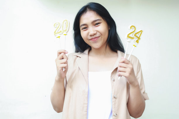 快乐的亚洲年轻女子手持金色数字蜡烛，微笑着对着镜头，穿着白色的休闲服装，庆祝2024年的除夕图片下载