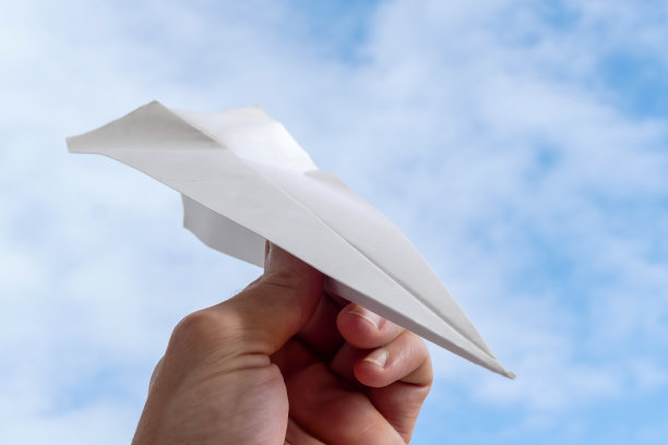 在蓝天白云的背景下，一个人手里拿着一架白纸飞机的近景图片下载