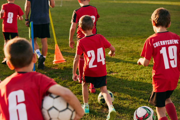 孩子们的后视图照片，穿着运动服准备训练的足球运动员，比赛前在足球场上拿着球。图片下载