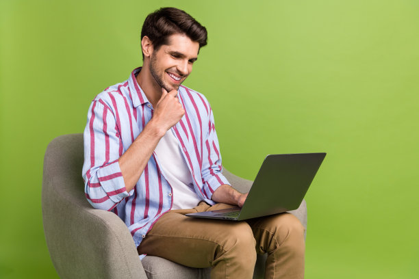 照片积极聪明的男人留着胡茬，穿着条纹衬衫，看着笔记本电脑思考，坐在扶手椅上孤立的绿色背景图片下载