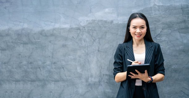 亚洲女商人身着黑色西装，手持数码平板电脑站在办公楼里，代表着以商业为导向的理念。商业库存照片。图片下载
