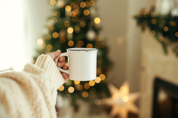 手持时尚的白色杯子与时尚的圣诞树，金色的灯和舒适的壁炉。杯子模型，圣诞广告产品模板。文本空间。冬天hygge图片下载