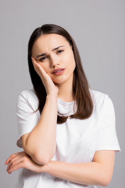 疲惫不堪的年轻女孩女士站在工作室的灰色背景上，孤立地望着远方，手靠近头，穿着基本的白色t恤，表现出不同的悲伤疲惫的情绪图片下载