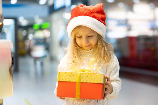 一个戴着圣诞帽、手里拿着新年礼物的小女孩站在玩具陈列柜前。孩子们为圣诞节挑选礼物。黑色星期五折扣图片下载
