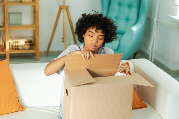 非洲女孩打开包裹，看着盒子。快乐的女人打开纸箱。女性拿着包裹，看着已经送达的物品。满意的客户积极反馈，快速快递交货。图片下载