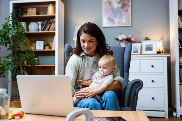 年轻的女企业主在休产假期间在家用笔记本电脑工作，抱着孩子经营私人公司。女商人，母亲，带着蹒跚学步的孩子在电脑前工作。图片下载