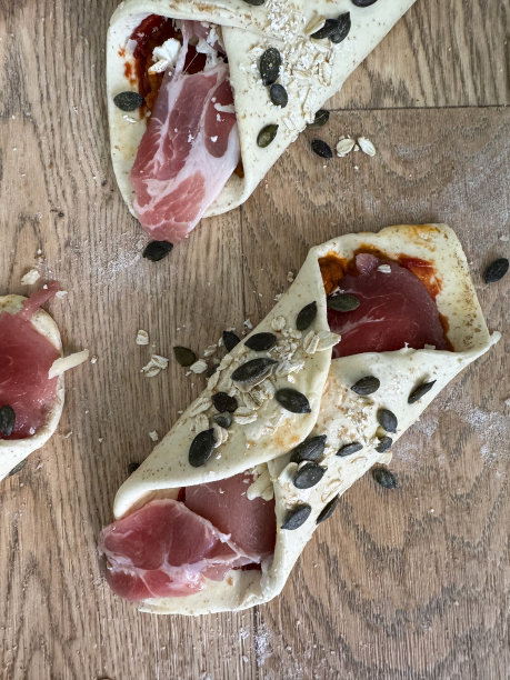 特写图片:未煮熟的、上了鸡蛋釉的、馅有番茄酱、培根和羊乳酪的油酥皮，上面撒着黑色葵花籽和燕麦，在撒了面粉的木纹背景上准备的图片下载