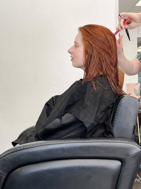 迷人的特写照片，红头发的女人坐在发廊的皮革扶手椅上，穿着黑色的理发师斗篷，无法辨认的人在剪湿头发，塑料红色梳子，理发师的剪刀，侧面视图，重点放在前景上图片下载