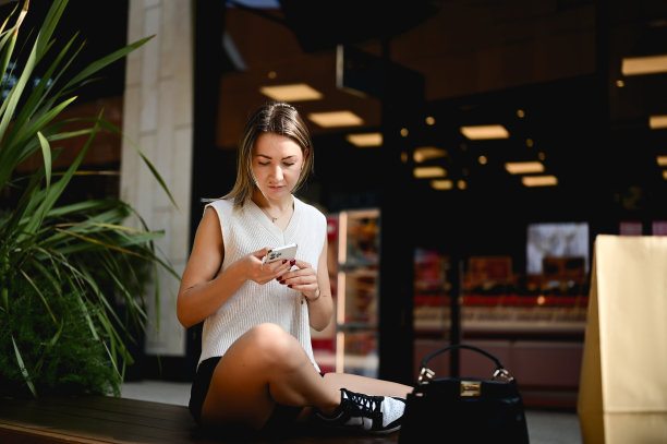 一个时髦的年轻女子坐在城市购物中心用智能手机网购的照片。图片下载