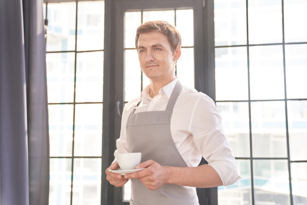 一位系着灰色围裙的男服务员端来一杯咖啡。咖啡师端上一杯热咖啡，背景是一扇大窗户，里面有可供复制的空间。高质量照片图片下载