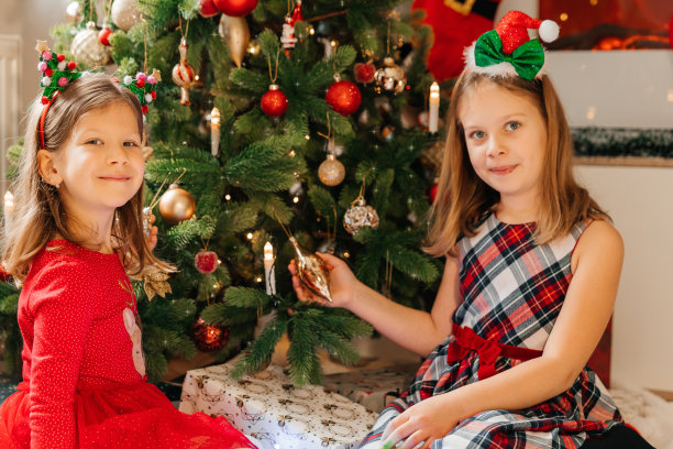 两个快乐的姐妹在新年树旁庆祝新年。孩子们装饰圣诞树。圣诞图片，树和灯的背景。圣诞快乐，新年快乐。图片下载