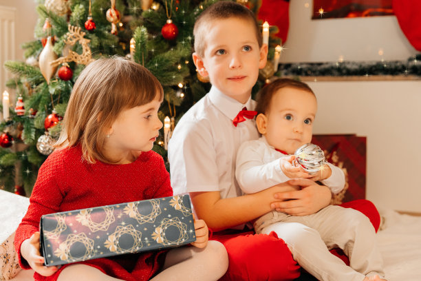孩子们在圣诞树旁打开新年礼物。哥哥和妹妹和一只狗坐在圣诞树旁边，享受着礼物。圣诞图片，树和灯的背景。圣诞快乐，新年快乐。图片下载