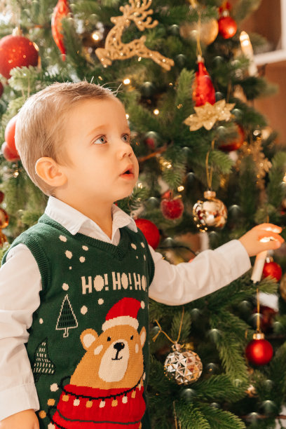 穿着节日服装的可爱小男孩站在圣诞树旁。圣诞快乐，新年快乐。圣诞图片，树和灯的背景。新年和圣诞节的概念。图片下载