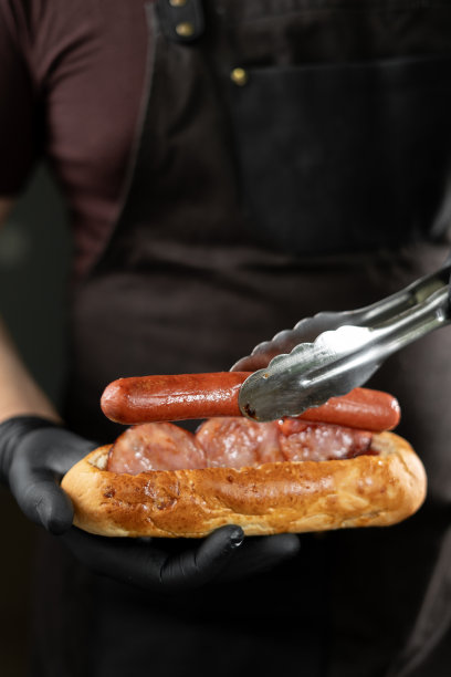 戴着黑色手套的厨师在做热狗，把香肠放在面包里。图片下载