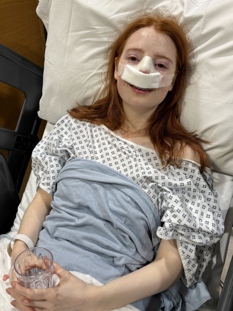 有吸引力的红发女人的特写照片，穿着医院的长袍，仰卧在康复室的床上，靠在枕头上，鼻中隔成形术后(鼻中隔偏曲手术和鼻子整形)，纱布绷带在鼻子上，拿着空杯子，聚焦于前景图片下载