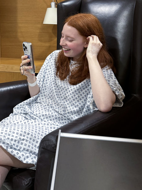 特写照片中，迷人的红发女性穿着医院的病号服，坐在医院候诊室的皮革扶手椅上，看着智能手机屏幕，鼻中隔成形术(鼻中隔偏曲手术和鼻子整形)，重点放在前景图片下载