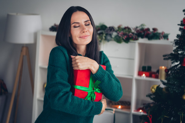 照片体贴甜美的女士穿着针织圣诞毛衣，闭上眼睛拥抱礼物在室内的房子房间图片下载