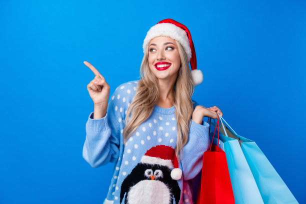 照片中可爱的金发女士手持购物袋点看空空间穿企鹅毛衣圣诞帽孤立的蓝色背景图片下载