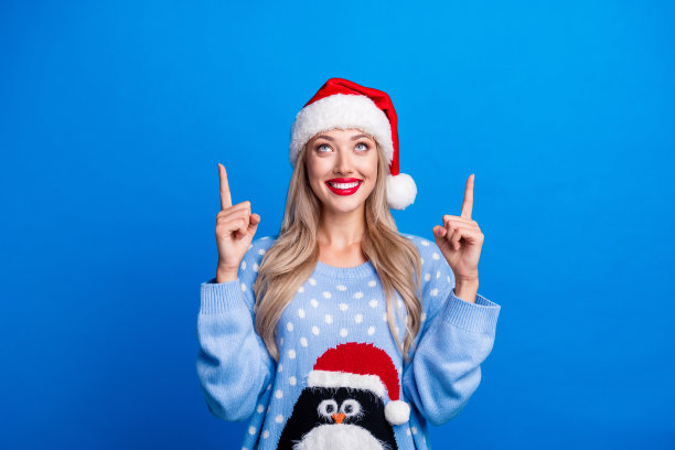 可爱的金发女郎的照片肖像指向看空空间穿企鹅毛衣圣诞帽孤立的蓝色背景图片下载