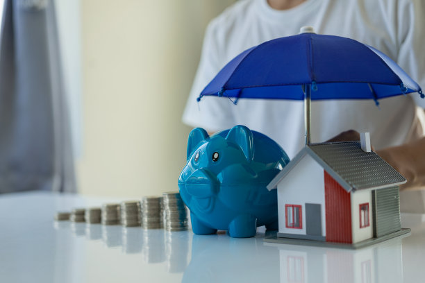 蓝色雨伞覆盖的房子模型和储钱罐在木制办公桌上与复印空间。经营房地产、财产保险、抵押保险的概念。图片下载