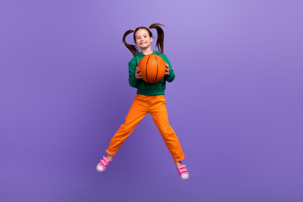全身照片甜美闪亮的小女孩穿着绿色卫衣跳高接篮球孤立的紫色背景图片下载