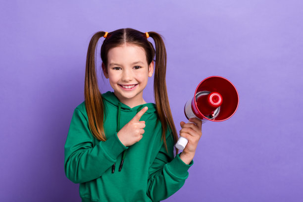 照片正面好心情的小女孩穿着绿色连帽衫，手指指向扩音器孤立的紫色背景图片下载