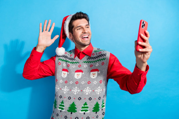 漂亮的年轻男性的照片肖像自拍挥手视频电话穿着圣诞印花背心帽子服装孤立的蓝色背景图片下载