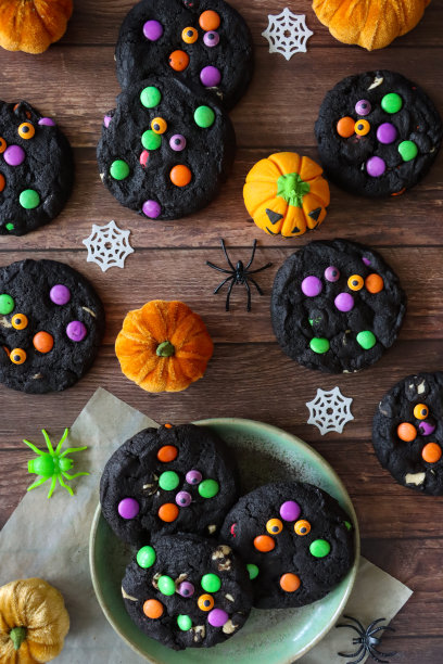 自制万圣节的特写图片，白色巧克力片，盘子上的黑可可饼干，装饰着粉红色，绿色和橙色的糖果涂层巧克力糖果，塑料蜘蛛，蜘蛛网，毛绒南瓜，木纹背景，高架视图图片下载
