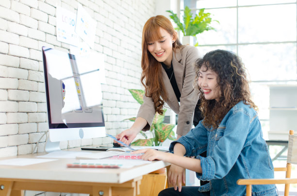 亚洲职业成功年轻女性创意平面设计师和同事，穿着休闲时尚套装，微笑着在办公室的电脑显示器上帮助挑选样品和比较艺术品图片下载