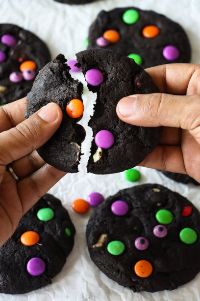 照片中，一个面目全非的人正在拍摄自制的万圣节，白色巧克力片，黑可可饼干，饼干批放在防油纸羊皮纸上，上面装饰着粉红色、绿色和橙色的巧克力糖果图片下载