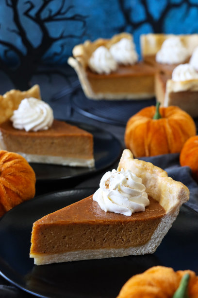 特写图片:黑色盘子上的一片新鲜出炉的万圣节南瓜派，上面有搅打过的奶油，毛绒南瓜，在冷却架上装满南瓜泥的有凹槽的挞皮，黑色纸树的剪影，斑驳的蓝色背景图片下载