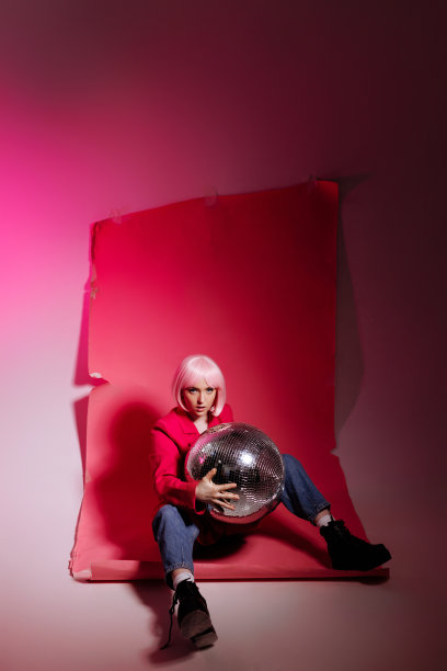 一名女模特坐在粉红色的背景上，拿着一个迪斯科球在摄影棚拍摄照片。图片下载