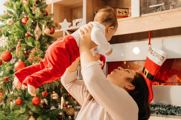 一位美丽的年轻母亲抱着她的小儿子，他们紧紧地拥抱在一起。母亲和她的孩子在圣诞树旁庆祝。圣诞图片，树和灯的背景。图片下载