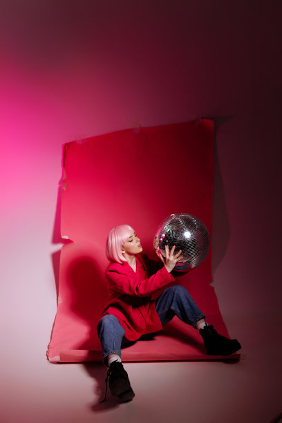一名女模特坐在粉红色的背景上，拿着一个迪斯科球在摄影棚拍摄照片。图片下载
