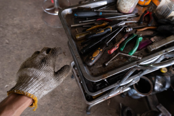 一个戴着安全手套的男人的左手正准备使用手动工具，螺丝刀、钳子和扳手来修理各种发动机。旁边还有一个工具架。图片下载