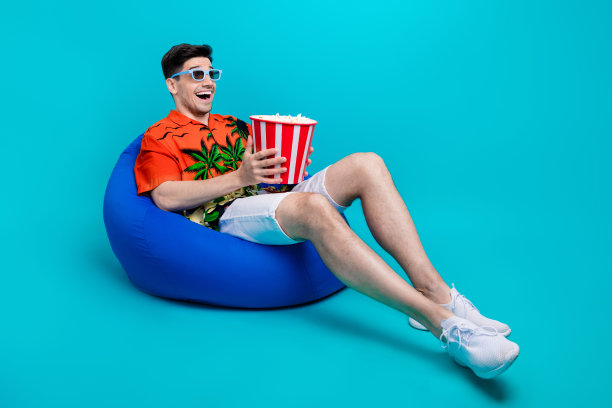 印象深刻的男人穿着印花衬衫的全尺寸照片爆米花在3d眼镜躺在pouf上看电影孤立的蓝色背景图片下载