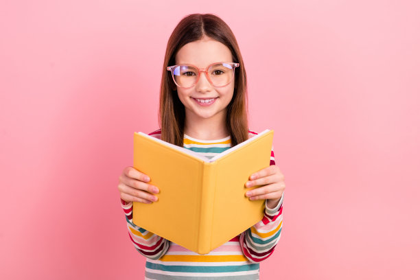乐观的小学习者的照片，十几岁前的女学生，戴着眼镜眼镜，在粉红色的背景上阅读抄写本图片下载