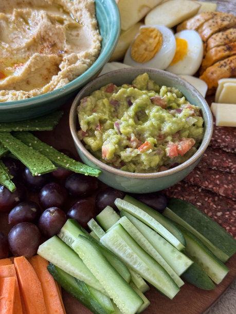 木制的、派对自助餐的全画幅图片，上面有几碗鳄梨酱和胡姆斯，周围是苹果、半熟的煮鸡蛋、香肠卷、切达奶酪、红葡萄、黄瓜、胡萝卜条和饼干图片下载