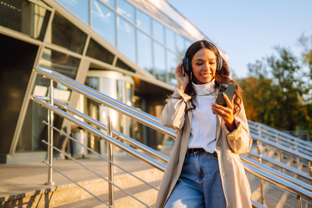 微笑的女人穿着时髦的衣服，拿着电话和无线耳机听音乐。图片下载