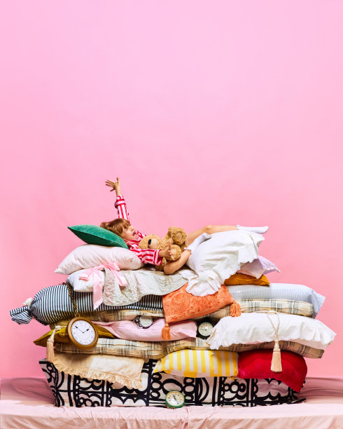 早上好。可爱的小公主和可爱的泰迪熊躺在大羽毛床上的肖像，粉红色的工作室背景上有枕头。图片下载