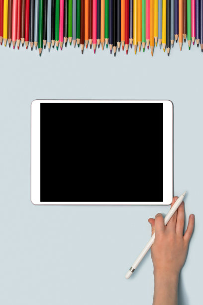 桌上放着一块写字板和一个拿着笔的孩子的手，上面放着五颜六色的铅笔。图片下载