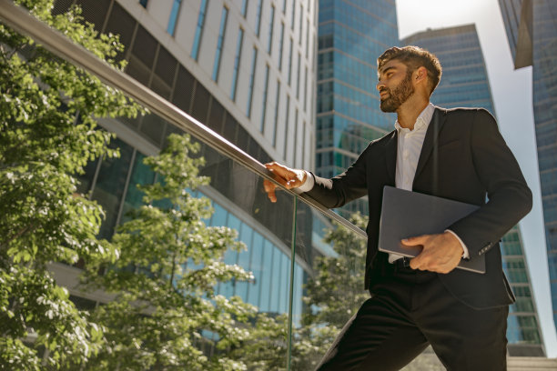 时髦的男经理拿着笔记本电脑站在办公室的阳台上，目光转向别处。高质量照片图片下载